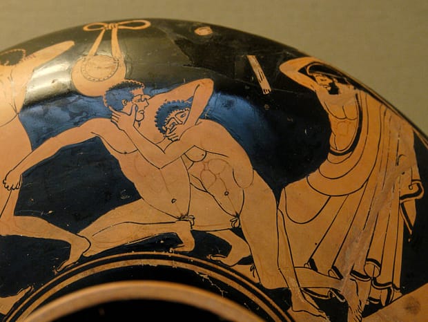 Red-figure cup painting of two pankration fighters grappling as one gouges an eye while the trainer raises a rod to stop the foul